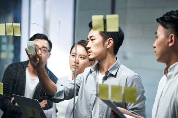 A group of people looking at sticky notes on a glass wall  Description automatically generated