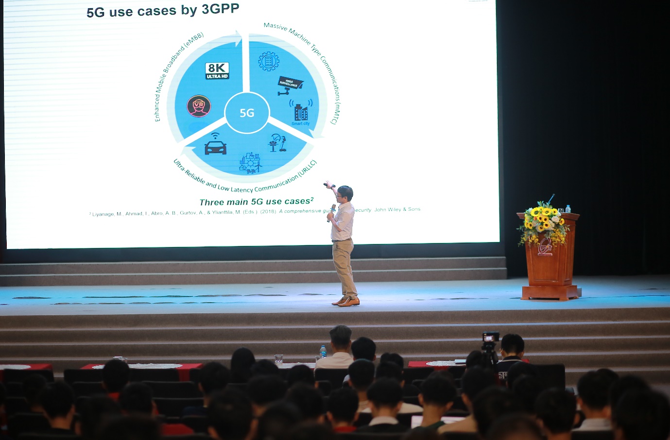 Những thách thức mới về an ninh mạng 5G cho hạ tầng số- Ảnh 2. A person standing on stage with a large screen Description automatically generated